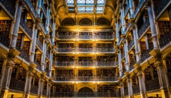 George Peabody Library Baltimore Visitor Guide Most visitors don't expect to find one of the most striking interiors in America tucked behind a row of white columns in a quiet Baltimore neighborhood. You walk through the doors of the Peabody Institute in Mount Vernon, turn a corner, and suddenly you're standing beneath a skylit atrium that climbs six stories above a polished marble floor. It stops you cold. The George Peabody Library Baltimore is that kind of place. It doesn't announce itself from the street. There's no flashy signage, no grand plaza. But step inside the stack room and you'll see why people call it a "cathedral of books." Over 300,000 volumes line the walls, and the space itself feels like something pulled from a 19th-century novel you half-remember reading. Johns Hopkins University runs the place as part of its Sheridan Libraries system. This isn’t a museum replica—it’s a working research collection. You can visit for free during public hours, which makes it one of the easiest cultural stops in Baltimore. Whether you’re chasing that perfect symmetrical shot up through the iron balconies or just want a quiet ten minutes surrounded by something genuinely beautiful, the library delivers without asking much of your time or your wallet. Cast-Iron Balconies And The Soaring Atrium The first thing that hits you is the scale. Five tiers of ornamental cast-iron balconies rise from the ground floor and climb 61 feet to a massive skylight that floods the room with natural light. The effect is vertical and dramatic, like standing inside a very elegant cage made of books and ironwork. Each balcony tier is lined with gold-and-black volumes shelved behind low railings, and the repeating geometric patterns of the iron railings create a visual rhythm that photographers obsess over. Point your camera straight up from the center of the floor and you'll get that iconic symmetrical shot that's all over travel feeds. The image almost looks digitally generated, but it's real, and it's been here since 1878. The marble floor adds to the atmosphere. Sound carries differently in here. Footsteps echo softly, and conversations drop to whispers without anyone being told. The reading room on the ground level sits just off the main atrium, offering a quieter space with wooden tables and the kind of warm, worn-in feeling that modern libraries rarely manage. The light keeps the space from feeling like a museum. On a clear afternoon, sunlight pours through the skylight and shifts across the iron railings and book spines, changing the room's character every hour. You could visit twice in the same day and walk away with completely different impressions. A Quick History Of The Landmark George Peabody, a financier who got his start in Baltimore before heading off to London, founded the Peabody Institute in 1857. He wanted to give something meaningful back to the city that launched his career—a free public library, a lecture series, a music conservatory, and an art gallery. The library building itself took its sweet time, finally opening up in 1878. Baltimore architect Edmund G. Lind teamed up with Nathaniel H. Morison, the institute's first provost, to design the interior. Lind focused on that dramatic stack room—he wanted it to feel grand enough to match Peabody's vision but still practical for researchers. Local craftsmen made the decorative cast-iron balconies, and people immediately noticed the design. It stood out as one of the most distinctive library interiors on the East Coast. The collection changed hands a few times. In 1966, the City of Baltimore took over and ran things through the Enoch Pratt Free Library. Then, in 1982, Johns Hopkins University stepped in, and the library became part of the Sheridan Libraries system. These days, the focus is on 18th- and 19th-century works—architecture, religion, science, geography, literature—with gems from folks like Edgar Allan Poe and Walt Whitman. George Peabody Library Baltimore