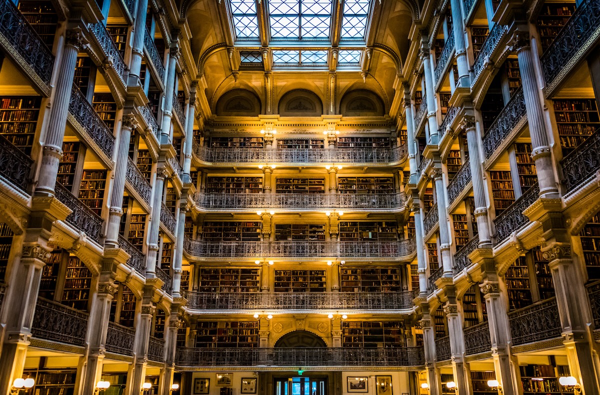 George Peabody Library Baltimore Visitor Guide Most visitors don't expect to find one of the most striking interiors in America tucked behind a row of white columns in a quiet Baltimore neighborhood. You walk through the doors of the Peabody Institute in Mount Vernon, turn a corner, and suddenly you're standing beneath a skylit atrium that climbs six stories above a polished marble floor. It stops you cold. The George Peabody Library Baltimore is that kind of place. It doesn't announce itself from the street. There's no flashy signage, no grand plaza. But step inside the stack room and you'll see why people call it a "cathedral of books." Over 300,000 volumes line the walls, and the space itself feels like something pulled from a 19th-century novel you half-remember reading. Johns Hopkins University runs the place as part of its Sheridan Libraries system. This isn’t a museum replica—it’s a working research collection. You can visit for free during public hours, which makes it one of the easiest cultural stops in Baltimore. Whether you’re chasing that perfect symmetrical shot up through the iron balconies or just want a quiet ten minutes surrounded by something genuinely beautiful, the library delivers without asking much of your time or your wallet. Cast-Iron Balconies And The Soaring Atrium The first thing that hits you is the scale. Five tiers of ornamental cast-iron balconies rise from the ground floor and climb 61 feet to a massive skylight that floods the room with natural light. The effect is vertical and dramatic, like standing inside a very elegant cage made of books and ironwork. Each balcony tier is lined with gold-and-black volumes shelved behind low railings, and the repeating geometric patterns of the iron railings create a visual rhythm that photographers obsess over. Point your camera straight up from the center of the floor and you'll get that iconic symmetrical shot that's all over travel feeds. The image almost looks digitally generated, but it's real, and it's been here since 1878. The marble floor adds to the atmosphere. Sound carries differently in here. Footsteps echo softly, and conversations drop to whispers without anyone being told. The reading room on the ground level sits just off the main atrium, offering a quieter space with wooden tables and the kind of warm, worn-in feeling that modern libraries rarely manage. The light keeps the space from feeling like a museum. On a clear afternoon, sunlight pours through the skylight and shifts across the iron railings and book spines, changing the room's character every hour. You could visit twice in the same day and walk away with completely different impressions. A Quick History Of The Landmark George Peabody, a financier who got his start in Baltimore before heading off to London, founded the Peabody Institute in 1857. He wanted to give something meaningful back to the city that launched his career—a free public library, a lecture series, a music conservatory, and an art gallery. The library building itself took its sweet time, finally opening up in 1878. Baltimore architect Edmund G. Lind teamed up with Nathaniel H. Morison, the institute's first provost, to design the interior. Lind focused on that dramatic stack room—he wanted it to feel grand enough to match Peabody's vision but still practical for researchers. Local craftsmen made the decorative cast-iron balconies, and people immediately noticed the design. It stood out as one of the most distinctive library interiors on the East Coast. The collection changed hands a few times. In 1966, the City of Baltimore took over and ran things through the Enoch Pratt Free Library. Then, in 1982, Johns Hopkins University stepped in, and the library became part of the Sheridan Libraries system. These days, the focus is on 18th- and 19th-century works—architecture, religion, science, geography, literature—with gems from folks like Edgar Allan Poe and Walt Whitman. George Peabody Library Baltimore