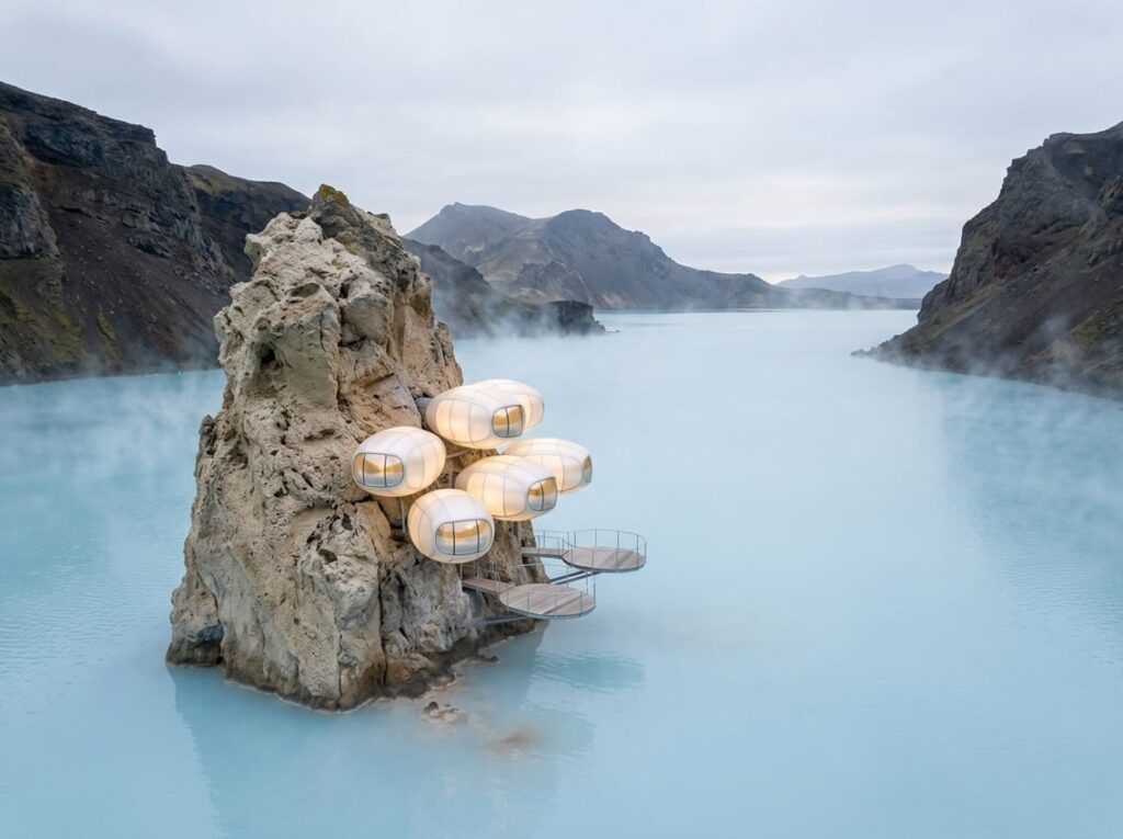 A cluster of glowing oval suites made of pearlescent panels anchored to a pale pumice sea stack in a volcanic bay with steaming milky blue water and suspended thermal soaking decks above the water.