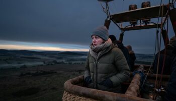 A bundled-up traveler stands in a hot-air balloon basket before sunrise, looking cold.